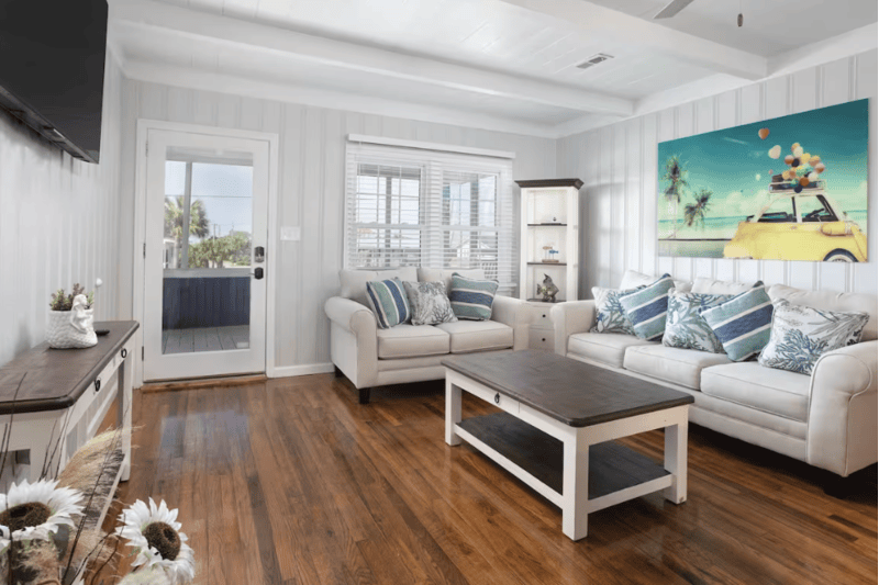 Sweet Home Carolina Bright living room with white sofas, wooden floor, beach-themed artwork, and large windows in a beach house.