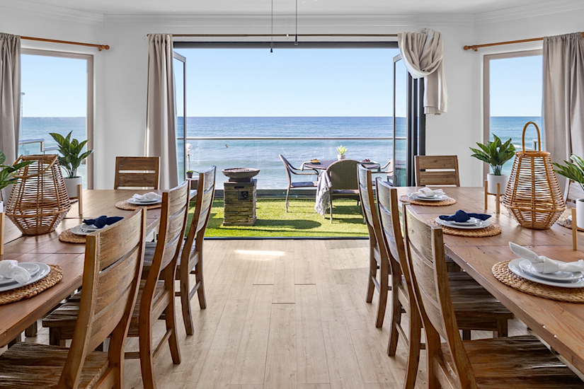 Bright dining room with large windows and glass doors opening to ocean view, beachfront balcony, and outdoor seating.