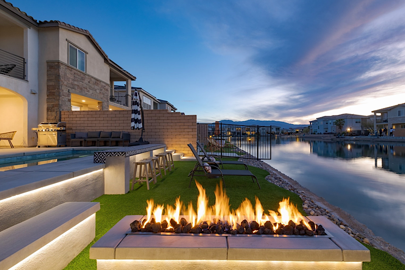 Coachella Valley Indio, CA Lago Luxe Luxury lakeside backyard in Indio, CA with a fire pit, pool, lounge chairs, and scenic mountain views at sunset.