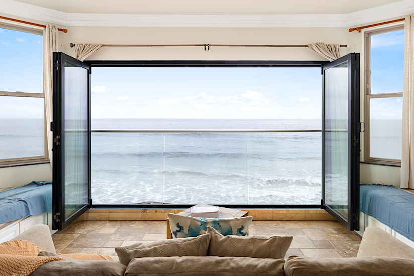 Living room with open glass doors overlooking the ocean, featuring built-in window seats and a cozy sofa, in a beachfront luxury home.