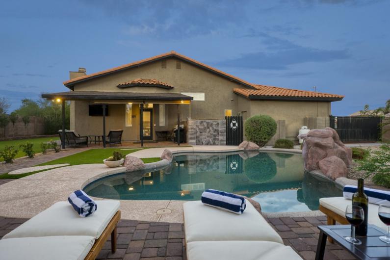 Luxury backyard with a swimming pool, lounge chairs, and a house, set against a backdrop of natural greenery and a blue sky.