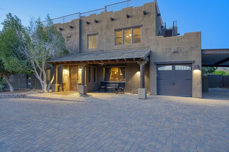 A modern, stucco-style house in Arizona with a spacious paved driveway, trees, and warm exterior lighting at dusk.