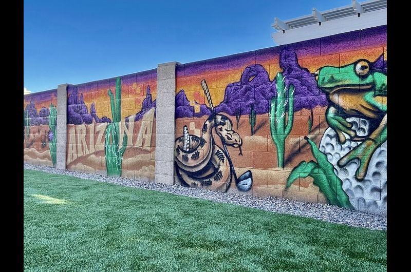 Colorful mural on a wall featuring desert landscape, cacti, a horse, and a frog, with a clear blue sky and green grass in front.