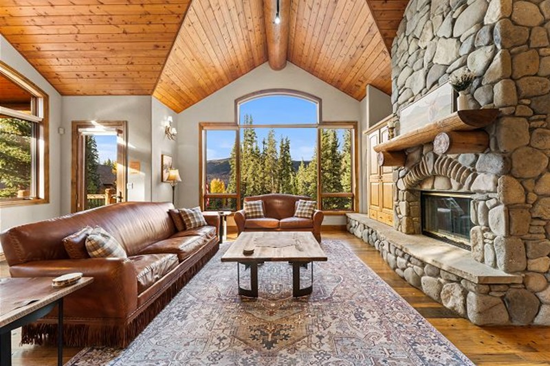 Cozy living room in a luxury mountain vacation home with a stone fireplace, wooden ceiling, large windows, and mountain views.