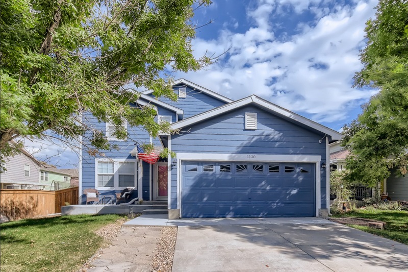 Bright blue 5-bedroom house in Brighton, Denver with a two-car garage, lush trees, and a well-maintained front yard.